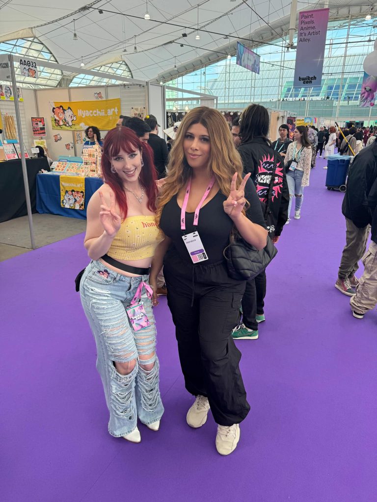 Two women posing at TwitchCon convention on purple carpet, smiling and making peace signs inside expo hall.